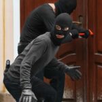 Due malviventi con viso coperto e vestiti di nero. Uno prova ad aprire una porta in legno con uno strumento rosso, l'altro fa da guardia e guarda dietro di sé